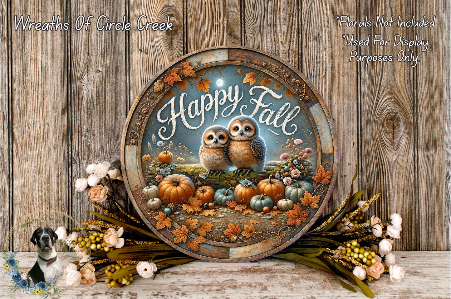 A decorative wreath with a pair of owls, pumpkins, and autumn leaves, surrounded by flowers and placed on a wooden surface.