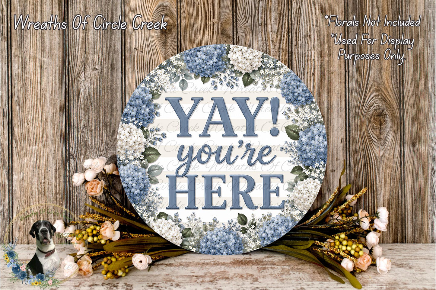 a round sign with the text "YAY! you're here" surrounded by a floral arrangement on a wooden surface.