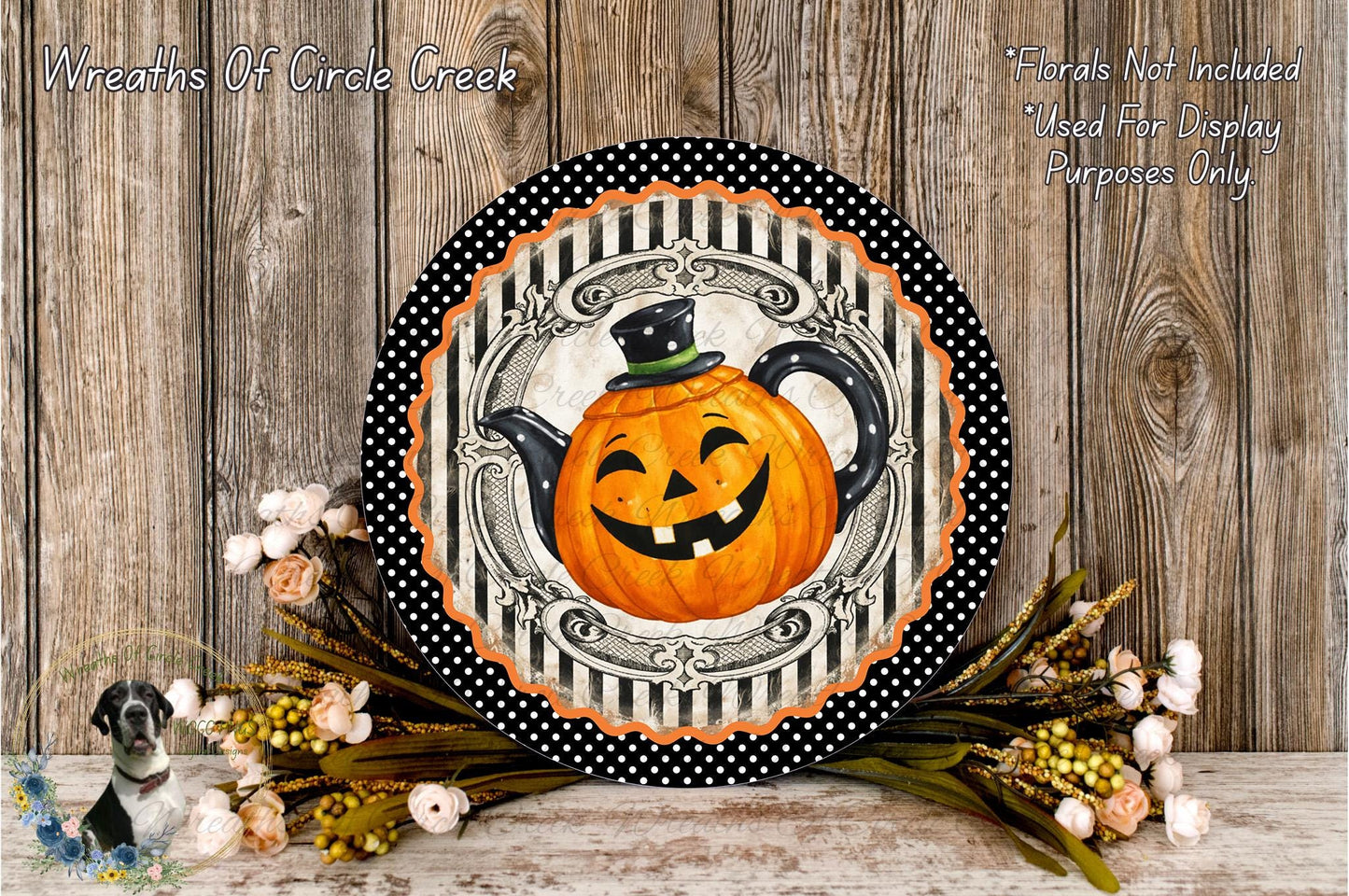 A Halloween-themed wreath with a smiling pumpkin teapot design, surrounded by flowers and placed on a wooden surface.