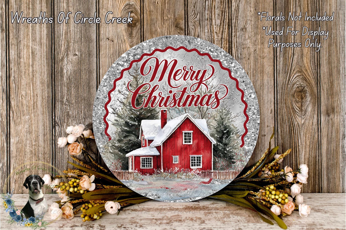 A round metal sign with a red house and snow-covered trees, surrounded by a wreath of flowers and greenery, against a wooden background.