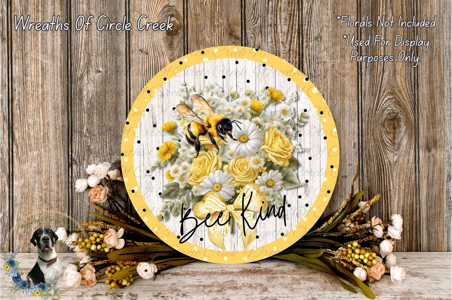 A decorative round sign with a floral design, featuring a bee and flowers, placed on a wooden surface. The sign has the text "Bee Kind" written on it.