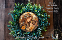 A wreath made of green leaves and flowers, with a circular medallion featuring a woman and a child in the center.
