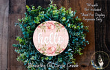 A wreath made of various green plants and flowers, with a pink circular sign displaying the word "Hello" in the center.