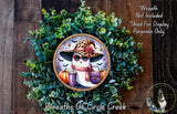 A wreath featuring a ghost wearing a leopard print hat, sunglasses, and holding a pumpkin and cup, surrounded by green foliage.