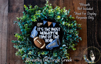 A wreath made of green foliage, with a football, cheerleading megaphone, and text that reads "It's the most wonderful time of the year" and "Go team!"