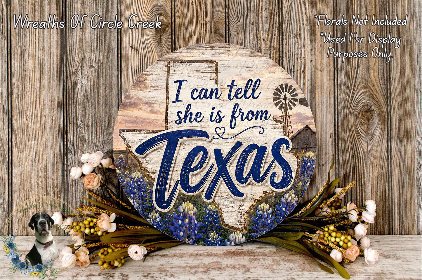 a round wooden sign with the text "I can tell she is from Texas" and a windmill illustration. The sign is surrounded by a wreath of flowers and placed on a wooden surface.