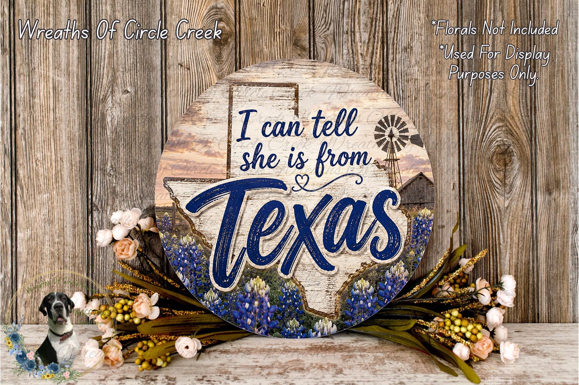 a round wooden sign with the text "I can tell she is from Texas" and a windmill illustration. The sign is surrounded by a wreath of flowers and placed on a wooden surface.