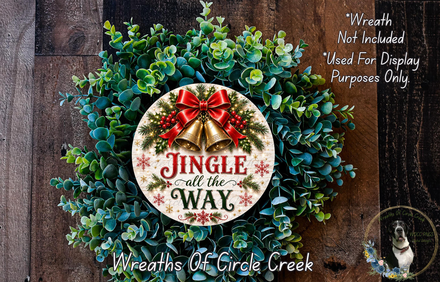 a Christmas wreath with a decorative sign that says "Jingle all the Way". The wreath is made of green leaves and has a red ribbon and bells attached to it. The sign is placed in the center of the wreath. The background of the image is a wooden surface with a dog visible in the bottom right corner.