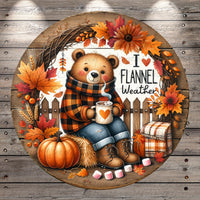 A round wooden sign featuring a teddy bear wearing a plaid sweater and holding a cup of hot chocolate, surrounded by autumn decorations such as a wreath, pumpkins, and leaves.