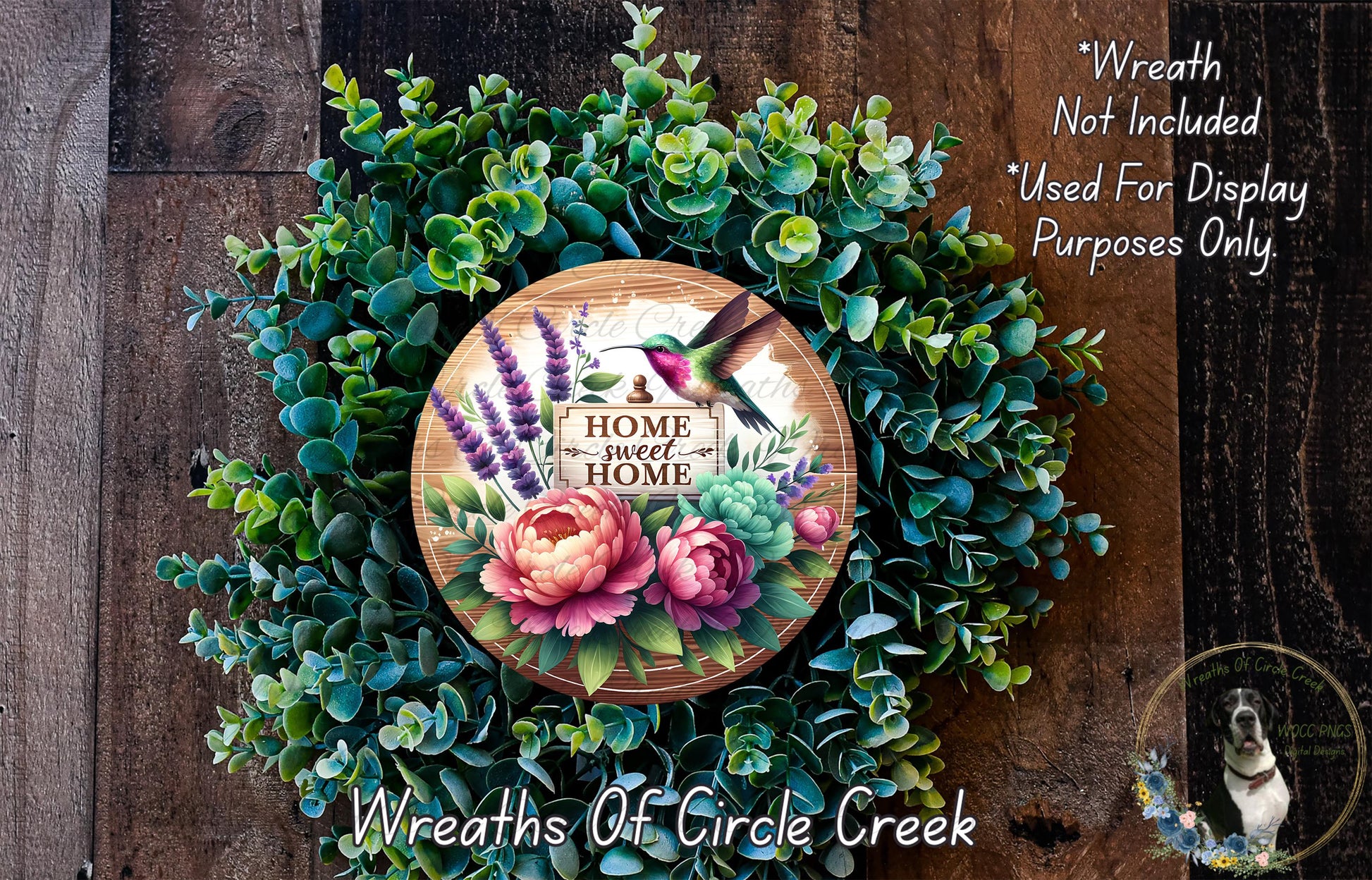 a wreath made of various green plants and flowers, with a wooden sign that reads "Home Sweet Home" in the center. The wreath is displayed on a wooden surface, and there is a small dog in the bottom right corner of the image.