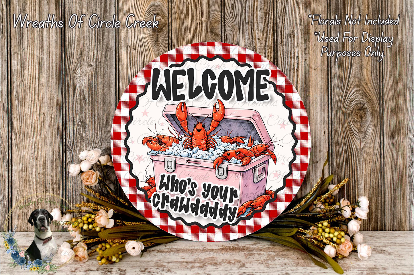 a circular sign with a red and white checkered border, displaying a cartoon lobster and the text "Welcome Who's Your Crawdaddy". The sign is surrounded by a wreath of flowers and placed on a wooden surface.