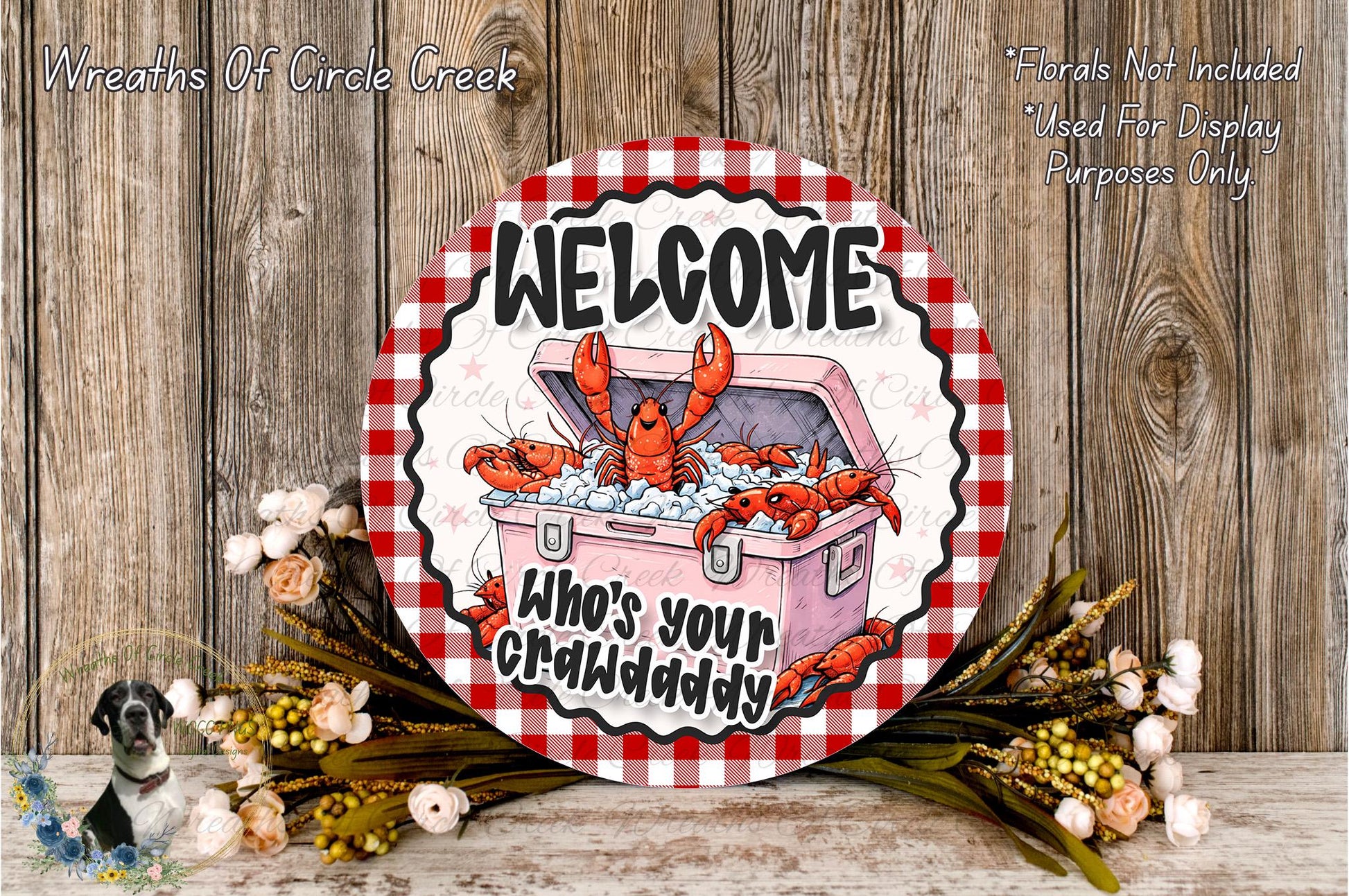 a circular sign with a red and white checkered border, displaying a cartoon lobster and the text "Welcome Who's Your Crawdaddy". The sign is surrounded by a wreath of flowers and placed on a wooden surface.