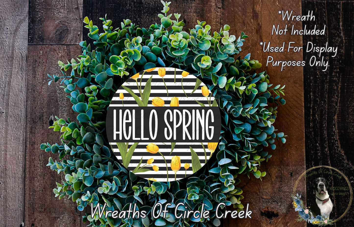 A wreath made of green and yellow flowers, with the text "Hello Spring" displayed on it. The wreath is placed on a wooden surface, and there is a small dog in the bottom right corner of the image.