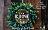 A wreath made of green and yellow flowers, with the text "Hello Spring" displayed on it. The wreath is placed on a wooden surface, and there is a small dog in the bottom right corner of the image.
