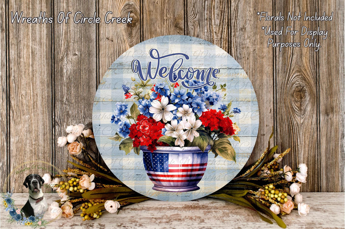 a round wooden sign with a patriotic design, displaying the word "Welcome" and a vase of red, white, and blue flowers. The sign is surrounded by a variety of flowers and foliage, creating a festive and welcoming atmosphere.