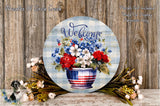 a round wooden sign with a patriotic design, displaying the word "Welcome" and a vase of red, white, and blue flowers. The sign is surrounded by a variety of flowers and foliage, creating a festive and welcoming atmosphere.