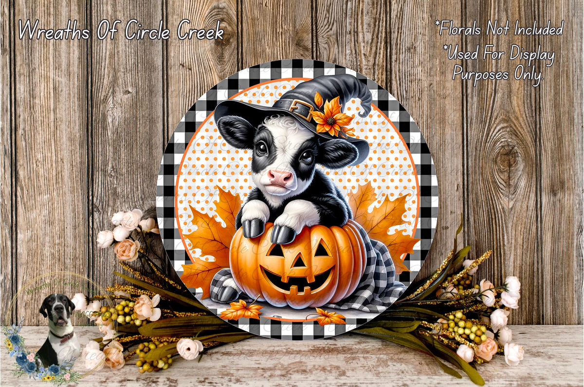 A black and white cow wearing a witch's hat is sitting on a carved pumpkin, surrounded by autumn leaves and flowers, with a wooden background.
