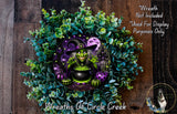 A wreath featuring a witch in a purple and green costume, surrounded by a lush green plant and a black pot with a green liquid inside.