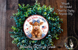 a wreath with a cow wearing a blue bow, surrounded by green foliage. The wreath is displayed on a wooden surface, and there is a small dog in the bottom right corner of the image.