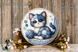A round wooden plaque with a cartoon cat sitting on a blanket, surrounded by flowers and a ball of yarn.