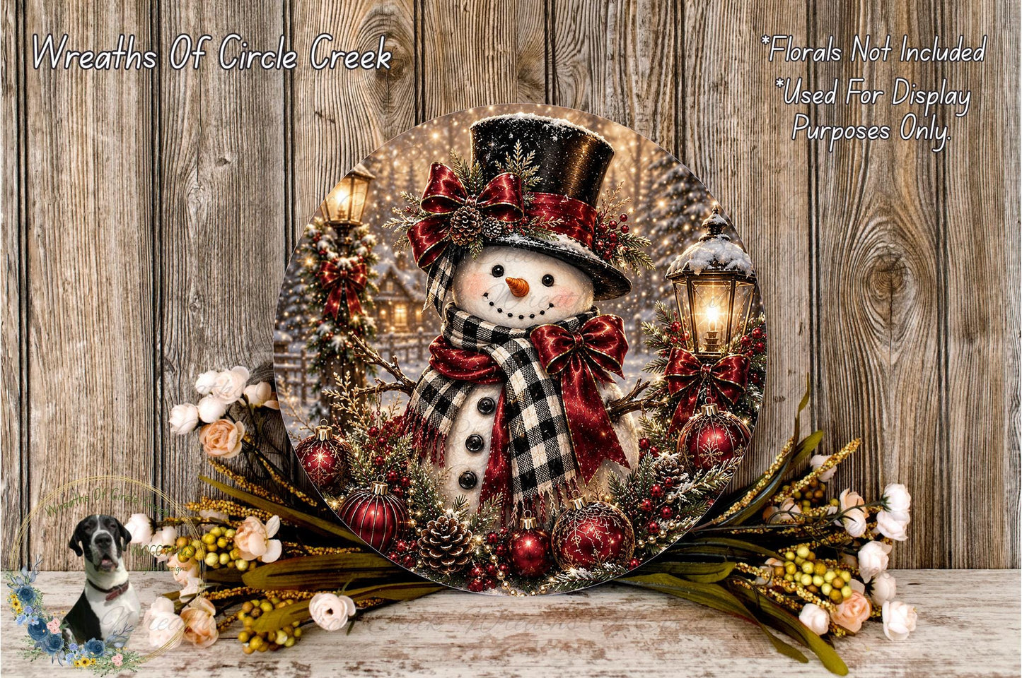 A snowman figure wearing a top hat and scarf, surrounded by a wreath of red and gold ornaments, pine cones, and flowers, set against a wooden background.