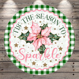 A circular decorative sign with a pink poinsettia flower, surrounded by green and white checkered patterns and gold stars. The sign has the text "Tis the Season to Sparkle" written on it.