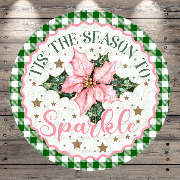 A circular decorative sign with a pink poinsettia flower, surrounded by green and white checkered patterns and gold stars. The sign has the text "Tis the Season to Sparkle" written on it.