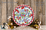 a decorative sign with the text "Home Sweet Home" surrounded by a red and white checkered border, placed on a wooden surface. The sign is surrounded by a floral arrangement, and there is a wooden wall in the background.