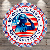 A circular design featuring a silhouette of a soldier saluting in front of an American flag, with the text "We don't know them all, but we owed them all" around the border.