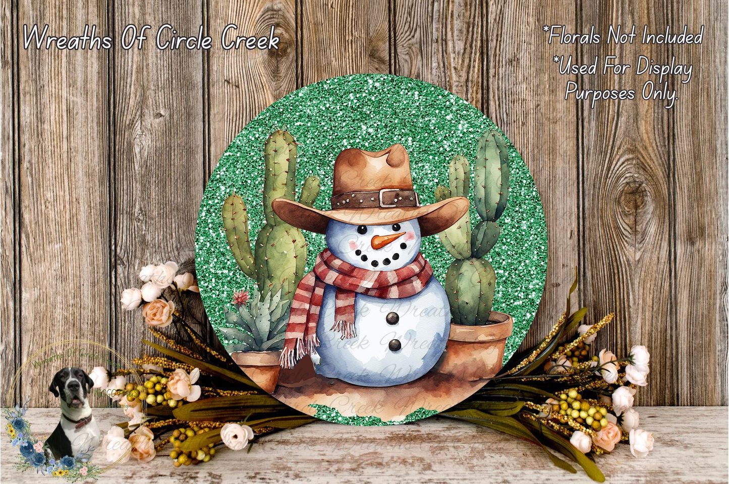 A snowman wearing a cowboy hat and scarf, surrounded by cacti and flowers, is displayed on a round, glittery plaque against a wooden background.
