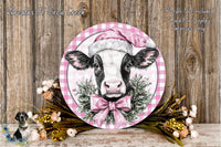 A decorative plate featuring a cow wearing a pink Santa hat, surrounded by a floral wreath and placed on a wooden surface.