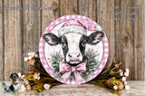 A decorative plate featuring a cow wearing a pink Santa hat, surrounded by a floral wreath and placed on a wooden surface.