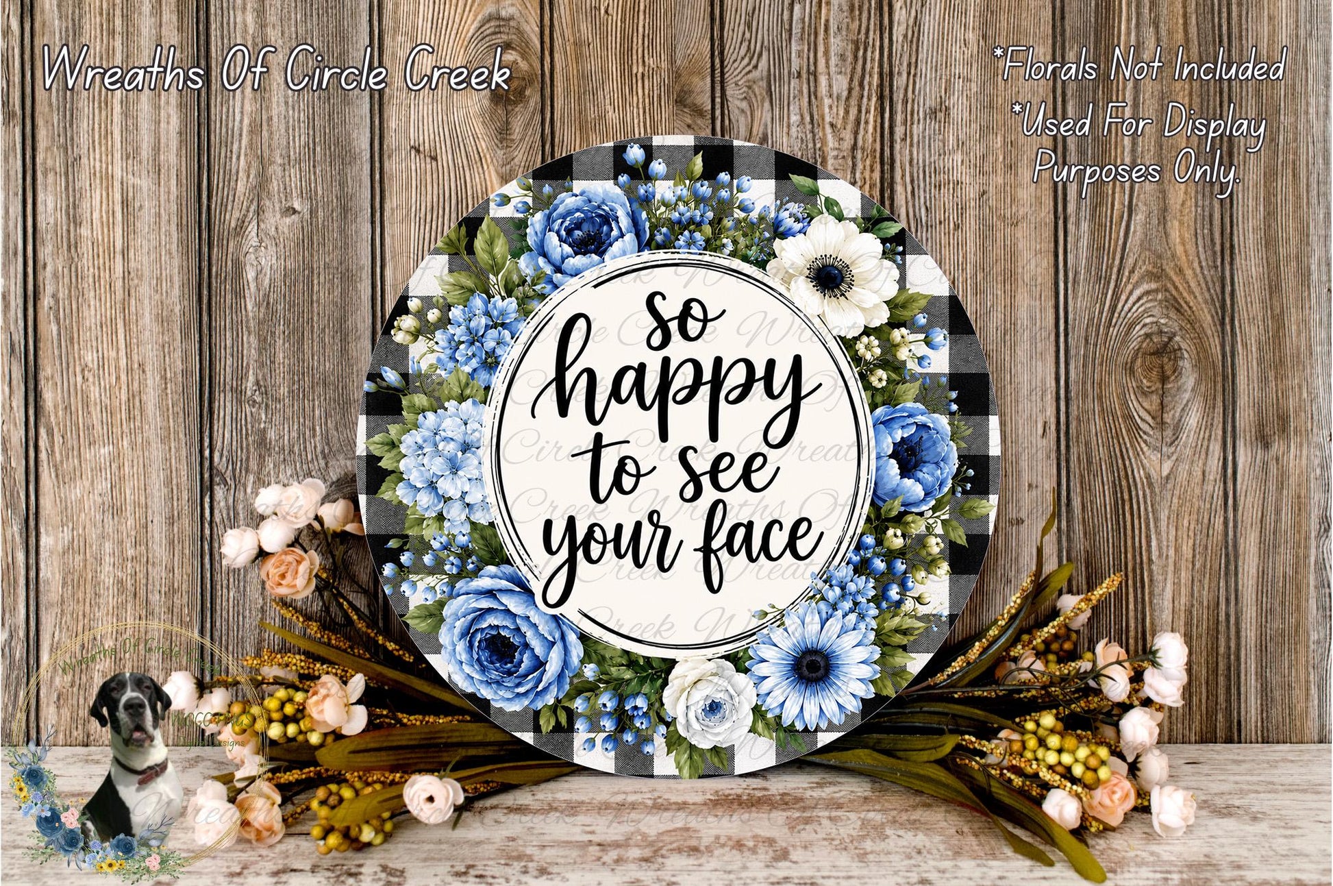 a decorative wreath with a floral design and the text "So happy to see your face" written on it. The wreath is surrounded by various flowers, including blue and white flowers, and is placed on a wooden surface.