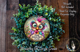 a wreath with a colorful owl design, surrounded by greenery and flowers. The wreath is placed on a wooden surface, and there is a small dog visible in the bottom right corner of the image.
