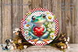 A round, decorative plate featuring a green frog holding a red heart, surrounded by a floral arrangement and placed on a wooden surface.