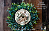 a wreath with a birdhouse design, surrounded by a lush green wreath. The wreath is placed on a wooden surface, and there is a small dog in the bottom right corner of the image.