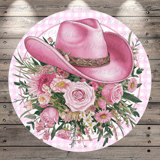 A pink cowboy hat adorned with a floral arrangement of pink and white flowers, including roses, daisies, and other blooms, is displayed on a circular background with a pink and white checkered pattern.