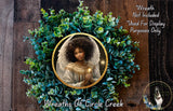 a wreath with a portrait of a woman, surrounded by green foliage. The wreath is displayed on a wooden background, and there is a small dog in the bottom right corner of the image.