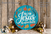 a blue circular sign with the text "FALL FOR JESUS he never leaves" surrounded by autumn leaves and flowers. The sign is placed on a wooden surface, and there are additional floral arrangements in the background.