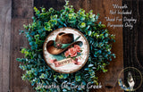 a wreath with a cowboy hat and the word "HOWDY" on it, surrounded by green foliage. The wreath is displayed on a wooden surface, and there is a small dog in the bottom right corner of the image.