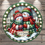 A festive winter scene featuring two cheerful snowmen, one wearing a green and red plaid outfit and the other in a red and green plaid outfit, standing together and smiling. The snowmen are surrounded by a wreath of berries and a sign that reads "Let it snow". The background includes a snowy landscape with a fence and trees covered in snow.