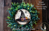 a wreath with a witch's hat and a spider web design, surrounded by green foliage. The wreath is displayed on a wooden surface, and there is a small dog in the bottom right corner of the image.