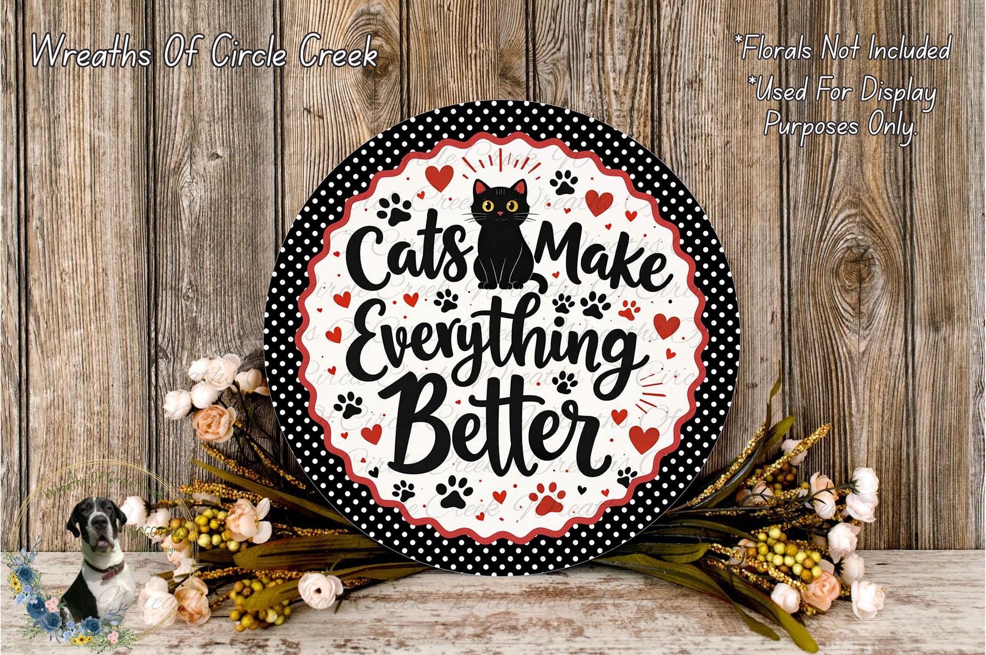 A decorative sign with the text "Cats Make Everything Better" surrounded by a black and white polka dot border, with a black cat illustration. The sign is placed on a wooden surface, and there are flowers and greenery in the foreground.