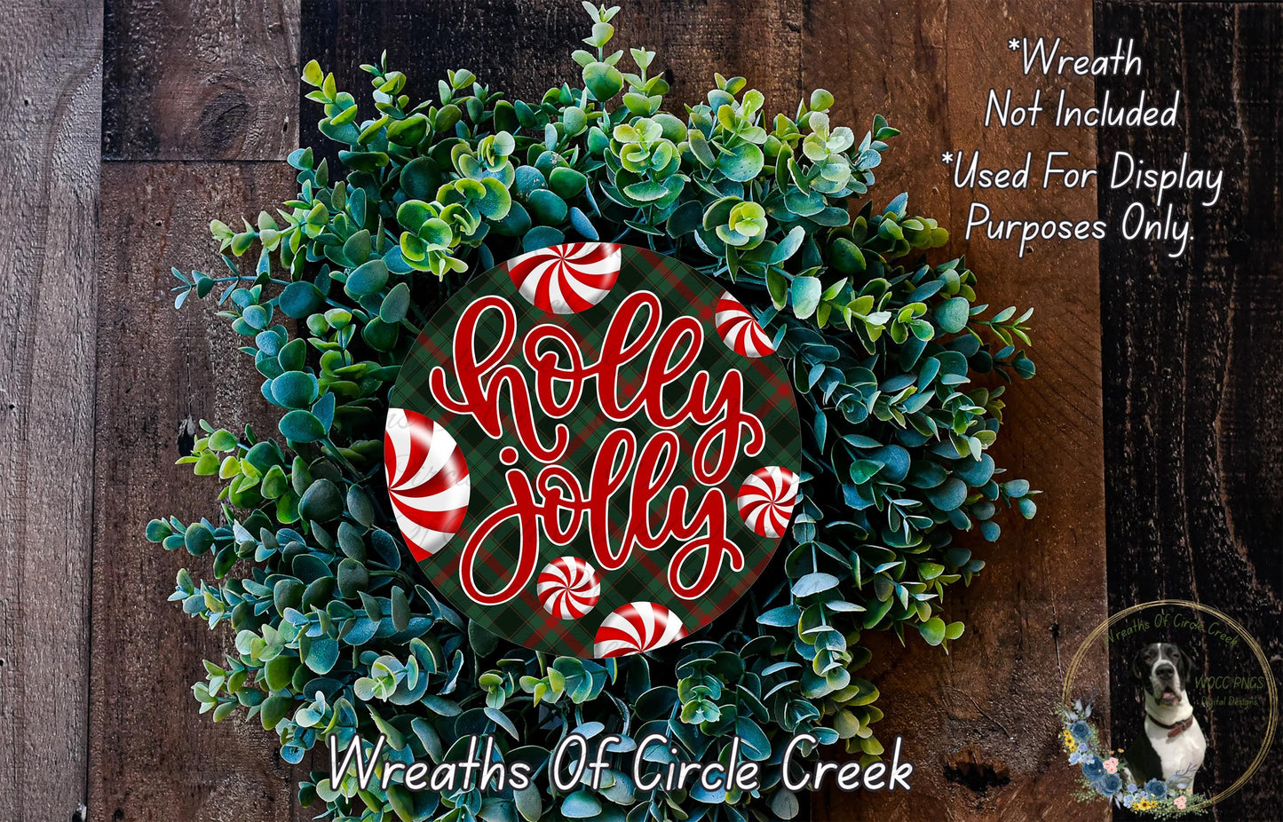 A wreath made of green and red leaves, with a red and white "Holly Jolly" sign in the center. The wreath is displayed on a wooden surface, and there is a small dog in the bottom right corner.