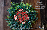 A wreath made of green and red leaves, with a red and white &quot;Holly Jolly&quot; sign in the center. The wreath is displayed on a wooden surface, and there is a small dog in the bottom right corner.