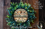 a wreath with a wooden sign that says "Welcome" surrounded by green foliage and white flowers. The wreath is placed on a wooden surface, and there is a small dog in the bottom right corner of the image.