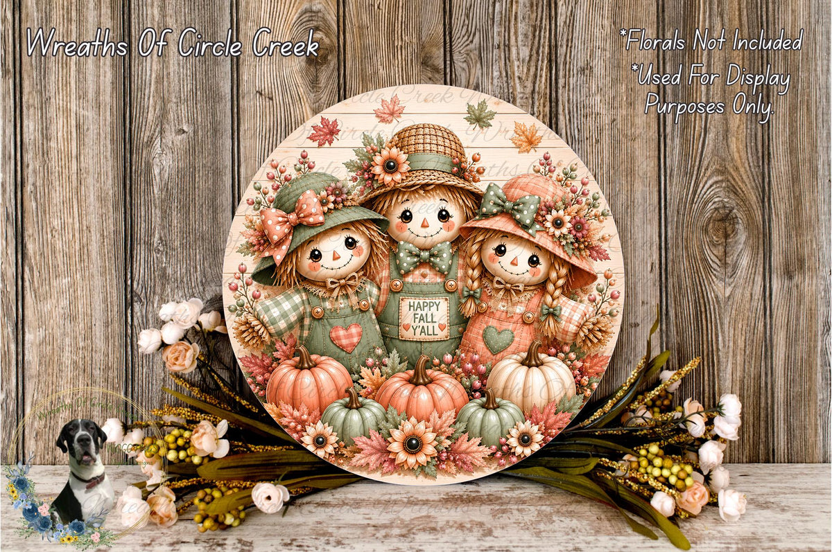 A round wooden sign with a cheerful autumn scene featuring three scarecrow dolls, pumpkins, and flowers. The sign is displayed on a wooden surface, surrounded by a floral arrangement.