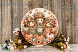 A round wooden sign with a cheerful autumn scene featuring three scarecrow dolls, pumpkins, and flowers. The sign is displayed on a wooden surface, surrounded by a floral arrangement.