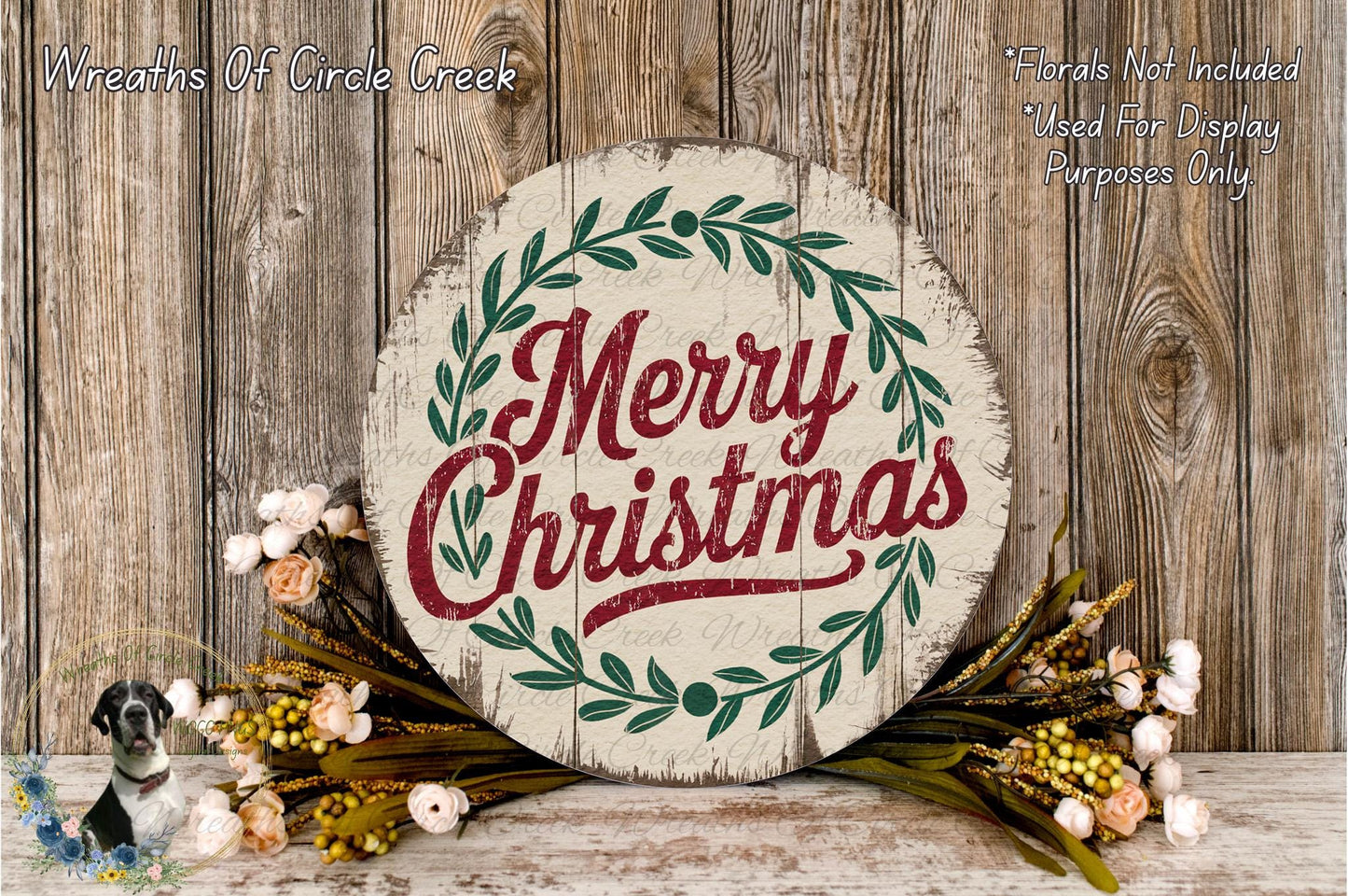 A round wooden sign with the text "Merry Christmas" in red and green lettering, surrounded by a wreath of greenery and flowers, placed on a wooden surface.