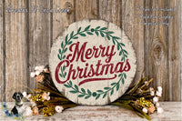 A round wooden sign with the text &quot;Merry Christmas&quot; in red and green lettering, surrounded by a wreath of greenery and flowers, placed on a wooden surface.